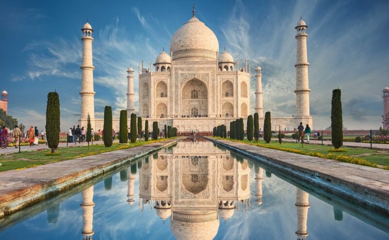 The Taj Mahal, a huge white marble building with domes and minaret towers against a blue cloud-streaked sky, with a still reflecting pool in front mirroring the building in it