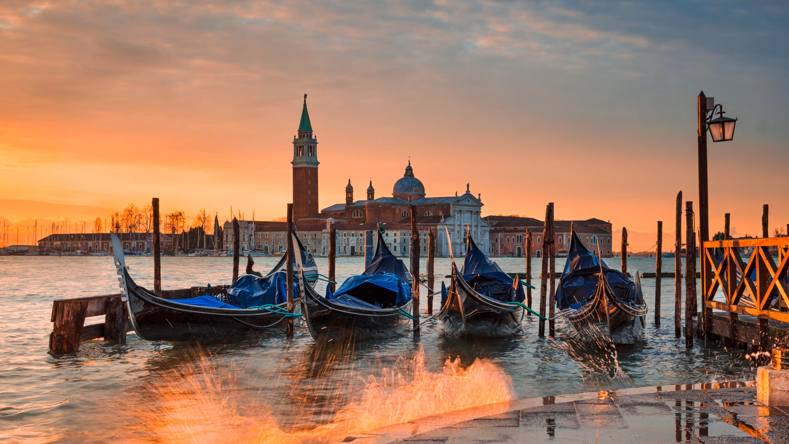 Gondolas docked at the water's edge in Venice during a bright orange sunset with a church tower in the background.