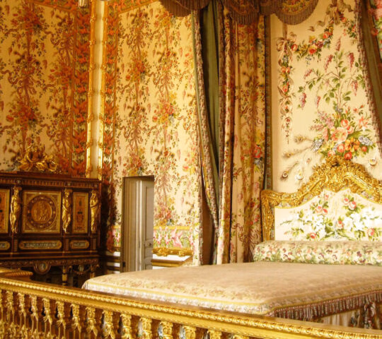 Ornate historical bedroom with colorful floral wallpaper, gold-framed bed, and green curtains with pink flowers.