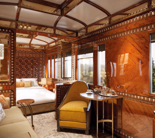 Interior of a luxury train cabin with high-gloss wood panels, a yellow chair, a table, and a bed in the background.