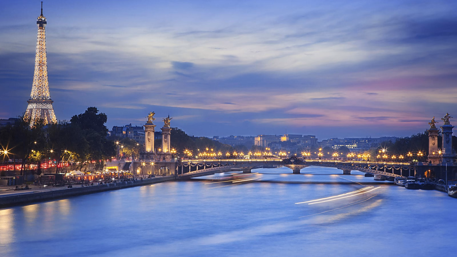 The Eiffel Tower at night beside the Seine River with a bridge and city lights reflected in the water.