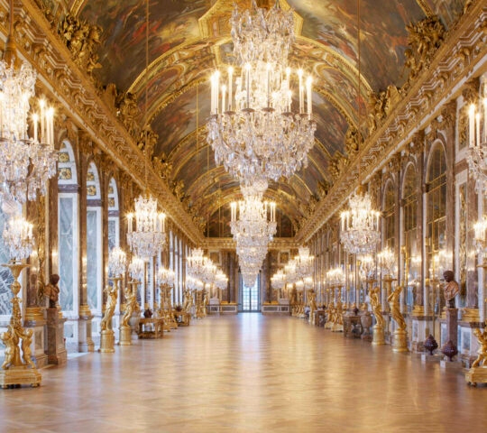 Grand interior of the Hall of Mirrors at Versailles with glowing chandeliers and ornate gold decor.
