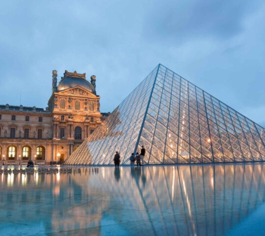The glass Louvre Pyramid in Paris at twilight, reflected in a still pool of water in front of the museum.