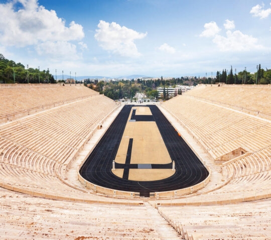 An empty marble stadium with a black running track under a blue sky with white clouds.