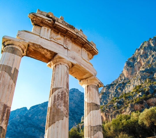 Ancient marble columns and ruins stand against a clear blue sky and a steep mountain side.