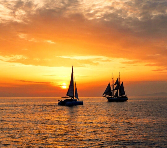 Silhouetted sailboats on the ocean under a bright orange and gold sunset sky with scattered clouds.