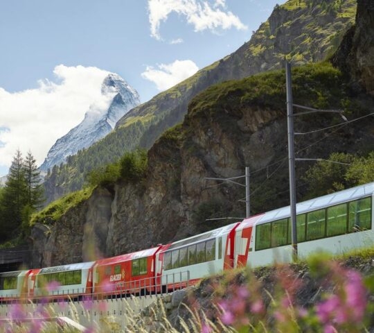 A red and white train traveling past a rocky cliffside with green trees and a mountain in the background.