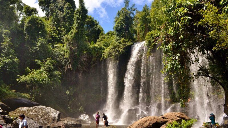 A scenic waterfall in a lush green forest with several people standing on rocks at the base of the falls.