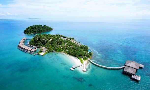 Aerial view of a tropical island resort with white sand, green trees, and bungalows over clear blue water.