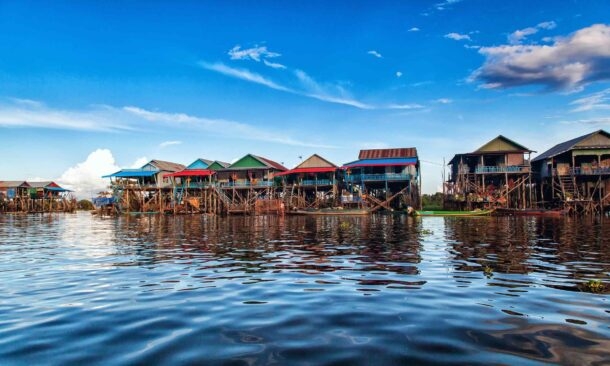 A row of colorful stilt houses in a floating village on a calm body of water reflecting the blue sky.