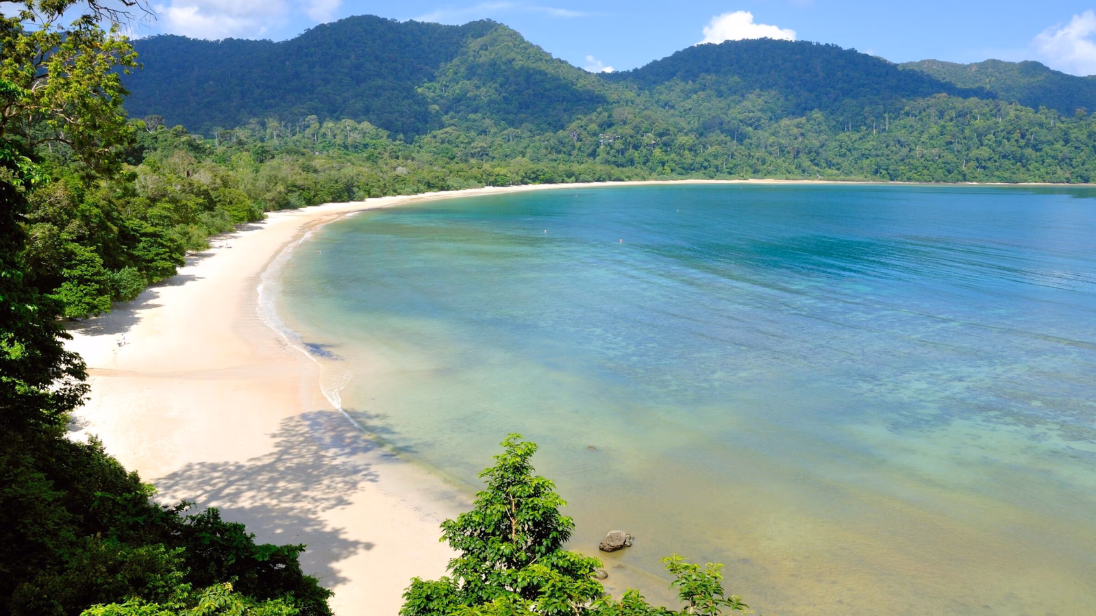 A serene tropical bay with a white sand beach and clear blue water surrounded by forested mountains.