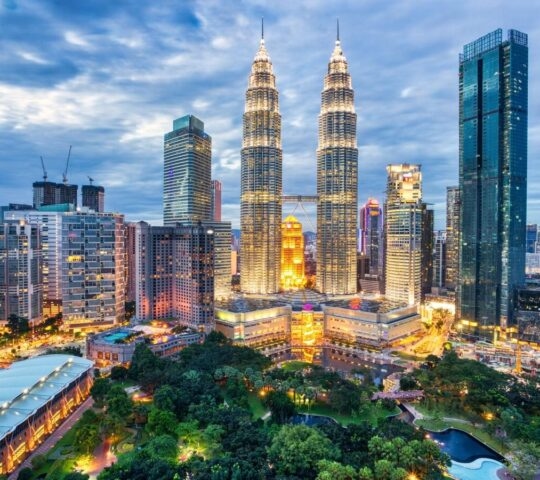 An aerial nighttime view of the illuminated Petronas Twin Towers and the surrounding city lights of Kuala Lumpur under a pink sky.