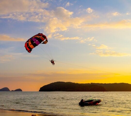A person parasails behind a boat over calm ocean waters against a vibrant orange and yellow sunset sky near an island.