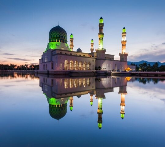 Large mosque with a green dome and minarets illuminated at dusk, reflected in clear water under a blue sky.