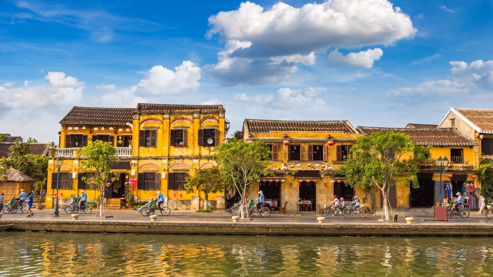 A row of bright yellow historic buildings along a riverbank with reflections in the water under a blue sky.