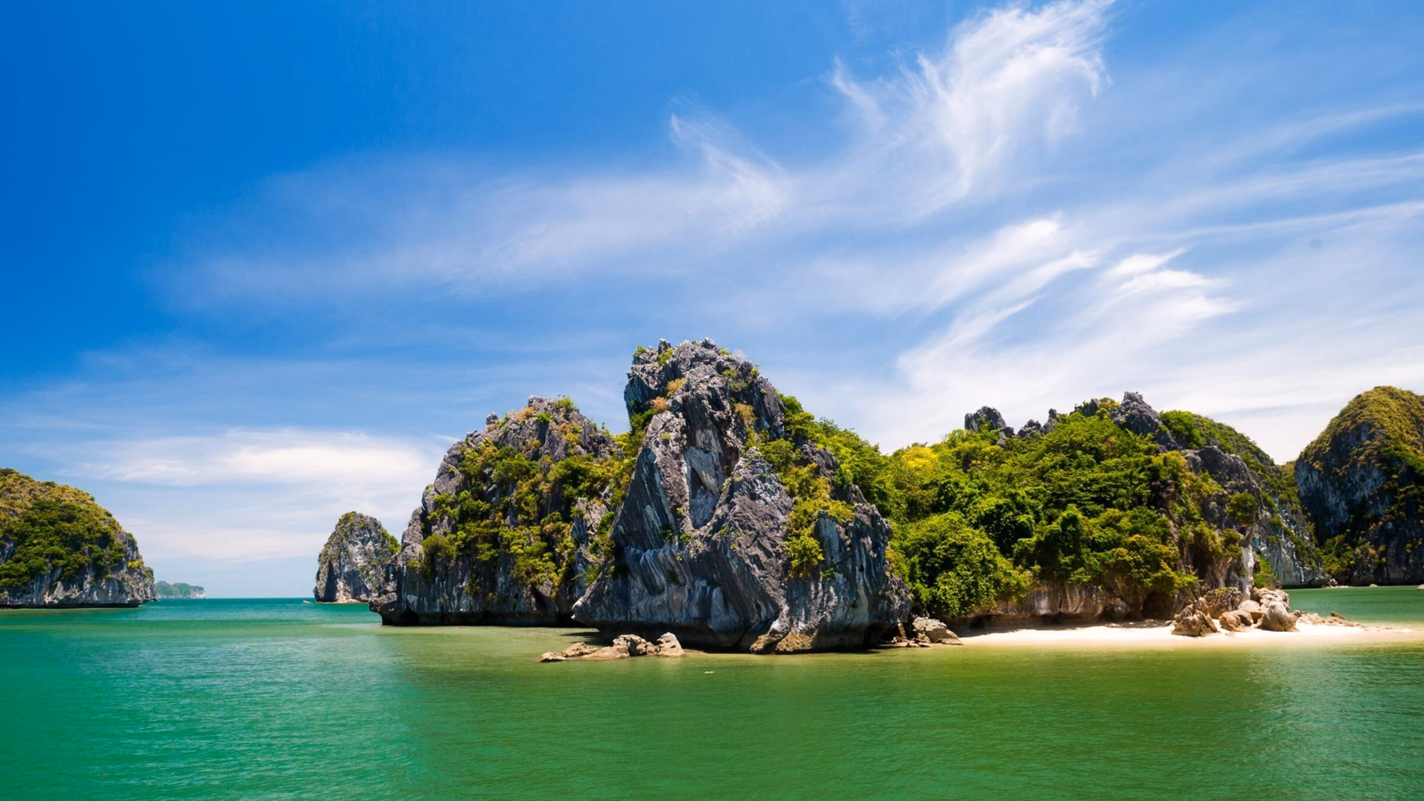 Steep, foliage-covered rock islands stand in clear green water under a blue sky with white clouds.
