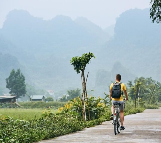 A man with a backpack cycles on a rural road toward distant, misty limestone mountains in a green landscape.