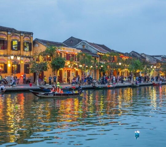 Illuminated yellow buildings and boats on a river at dusk with reflections shimmering on the water surface.