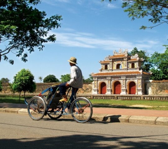 A man on a three-wheeled cycle taxi parked near an ancient decorative archway and green trees.