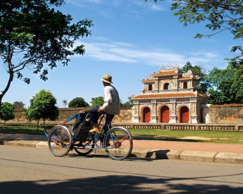 A man on a three-wheeled cycle taxi parked near an ancient decorative archway and green trees.
