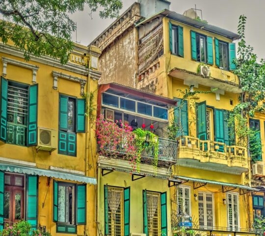 Close-up of yellow residential buildings with many teal-colored window shutters and a flower-filled balcony.