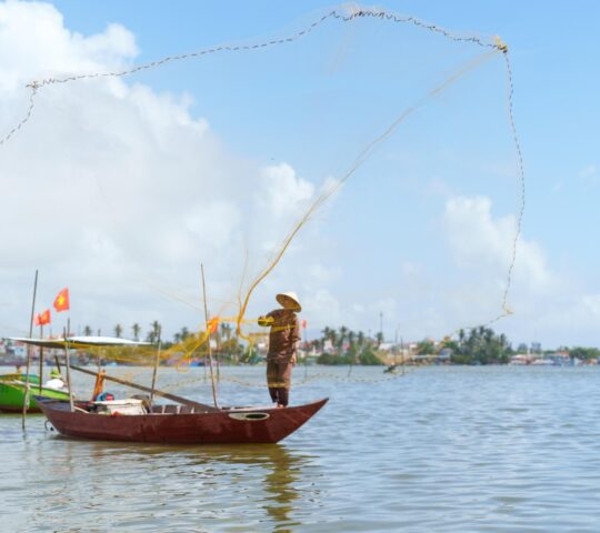A fisherman on a boat throwing a large fishing net into the air over the sea with a distant shoreline.