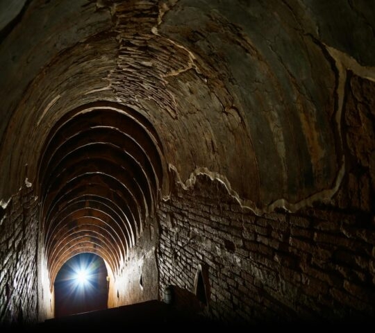 Perspective view inside a dark, arched brick tunnel with a bright light shining at the end of the corridor.