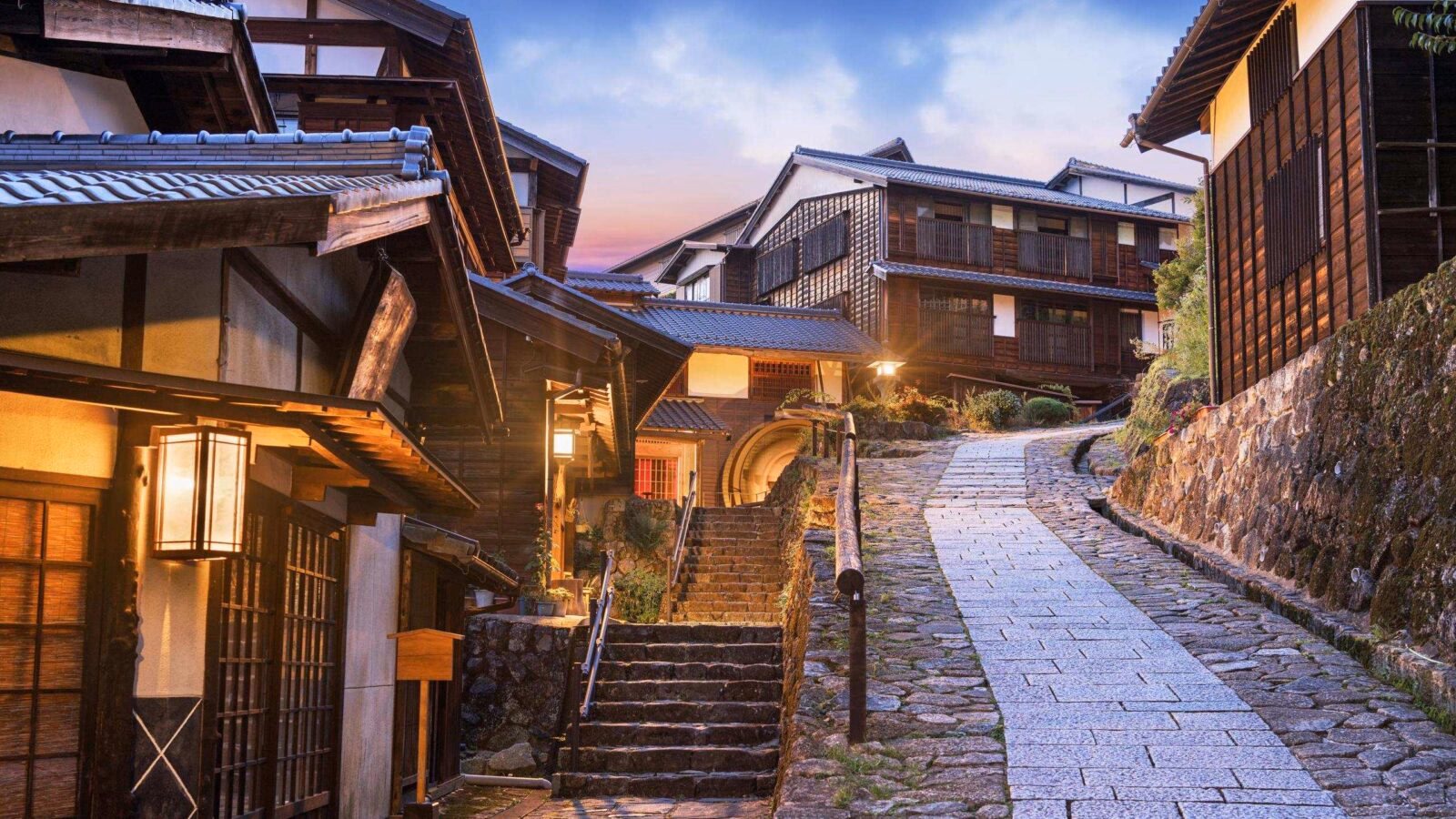 Stone-paved steps wind through a historic Japanese village with traditional wooden houses and glowing lanterns at dusk.