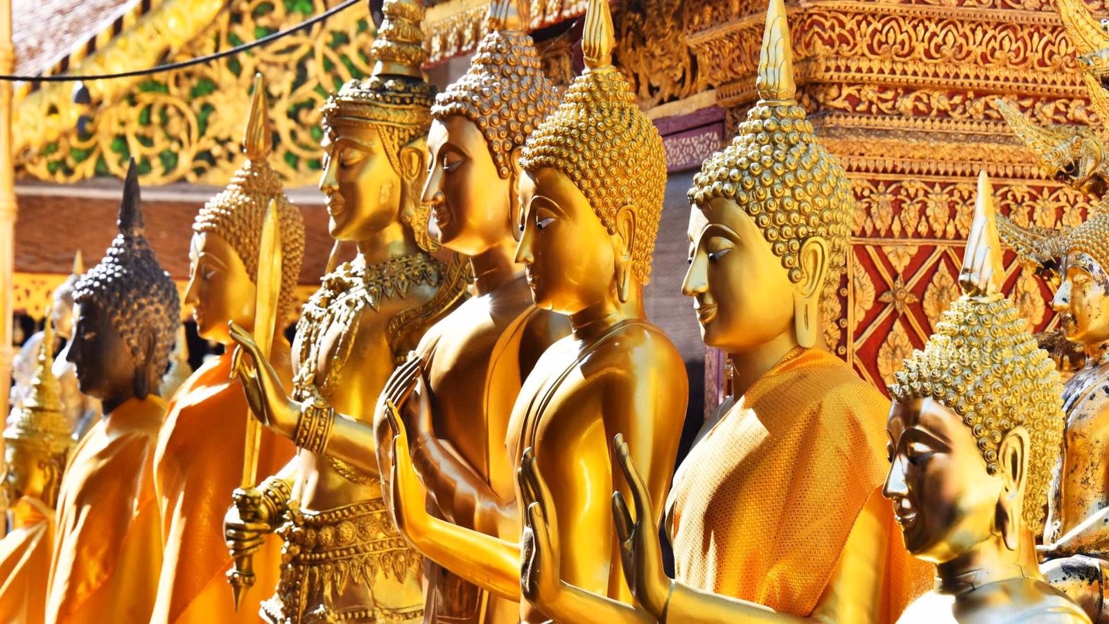 A close-up side view of several golden Buddha statues lined up in front of an intricately carved temple wall.