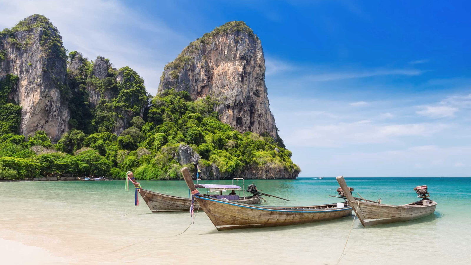 Three traditional wooden longtail boats moored on a sandy beach with large limestone cliffs and tropical green foliage.