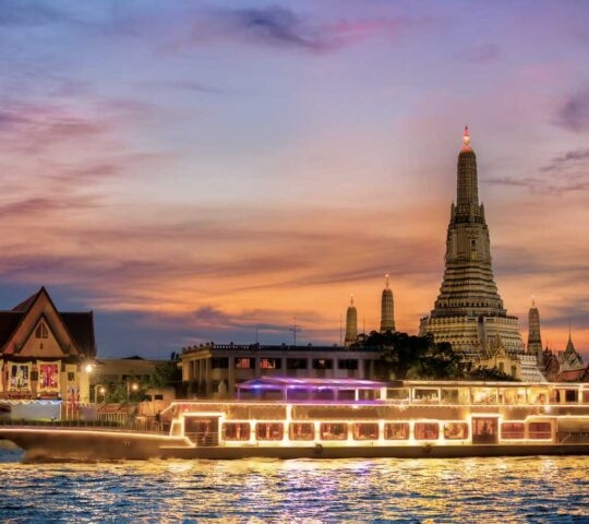 A cruise boat with lights reflecting on the water at night, with a tall, illuminated temple in the background.