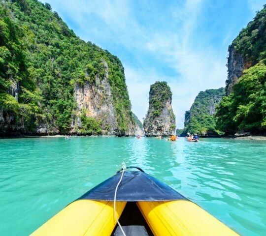 View from a yellow kayak on turquoise water between high, tree-covered cliffs under a bright blue sky.