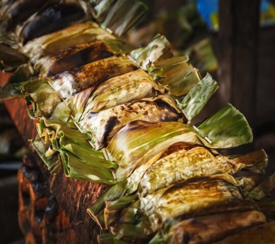 Close-up of street food wrapped in green leaves being grilled on a wooden rack.