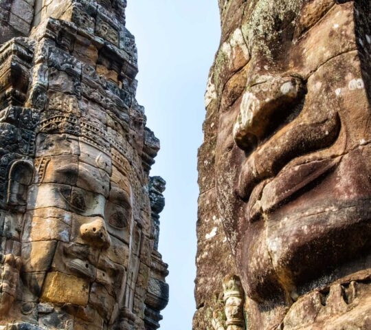Two large, weathered faces carved into ancient stone towers against a clear sky.