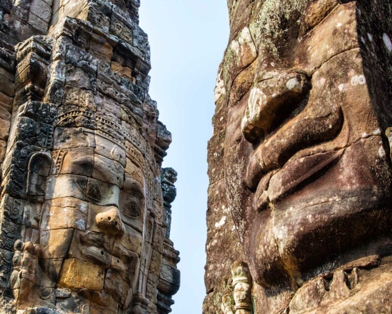Two large, weathered faces carved into ancient stone towers against a clear sky.