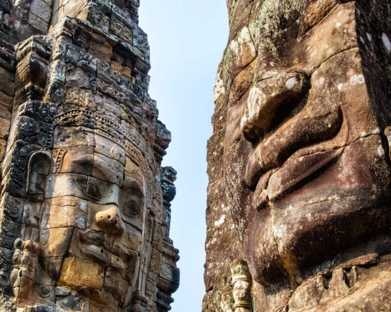 Two large, weathered faces carved into ancient stone towers against a clear sky.