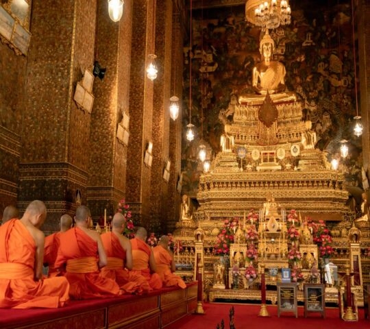 Buddhist monks in orange robes sit facing a large, ornate golden Buddha statue inside a temple.