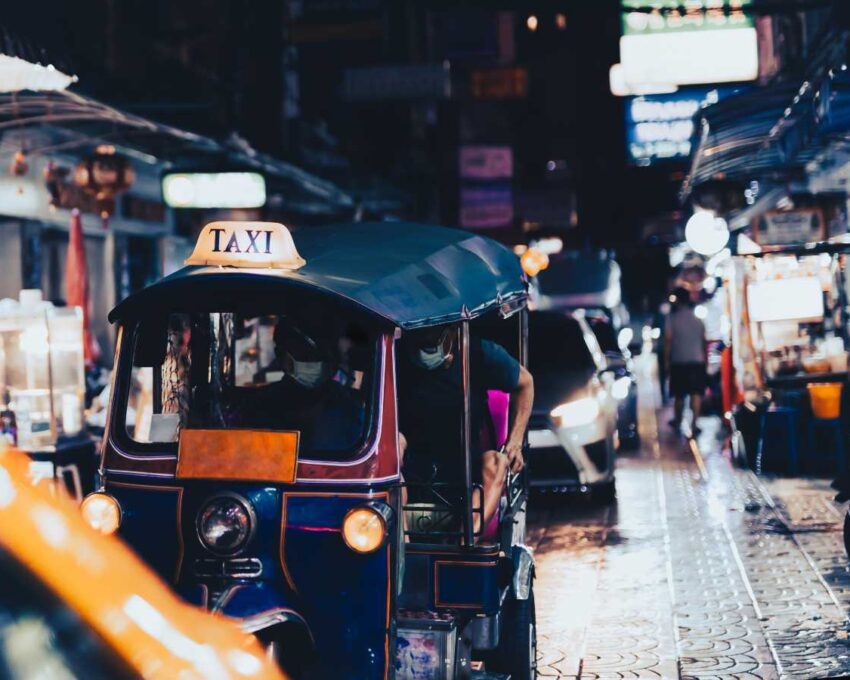 A blue and red tuk-tuk taxi with a yellow TAXI sign parked on a busy city street at night.