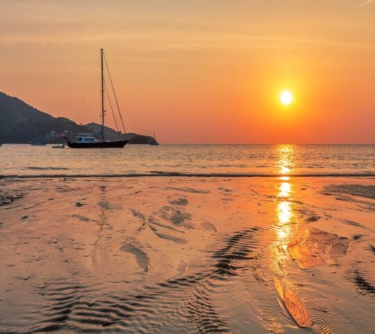 A sailboat anchored in a bay during a golden sunset with reflections on the wet shoreline.