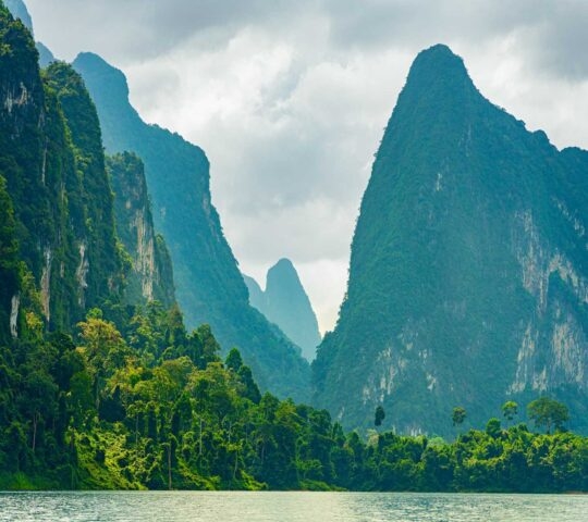 Lush green limestone karst mountains rising from a calm body of water under a cloudy sky.