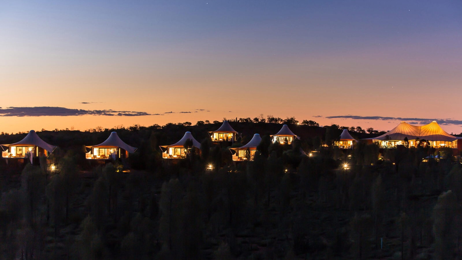 Several illuminated glamping tents on a ridge during a desert sunset with a colorful twilight sky.