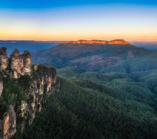 The Three Sisters rock formation overlooking a deep green valley in the Blue Mountains at sunset.