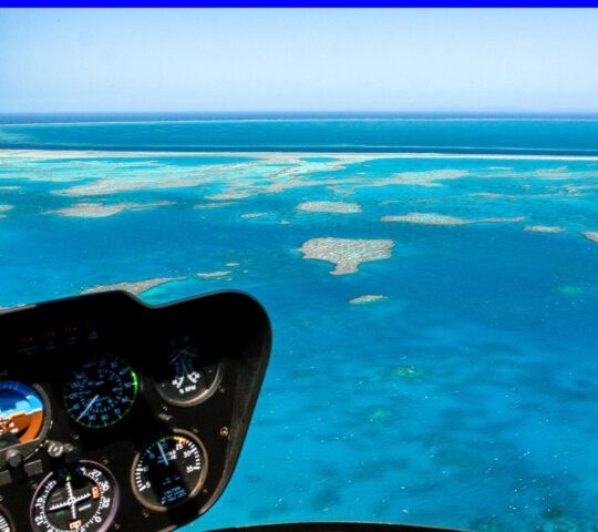 View from a helicopter cockpit showing instrument dials and a vast turquoise coral reef stretching toward the horizon.