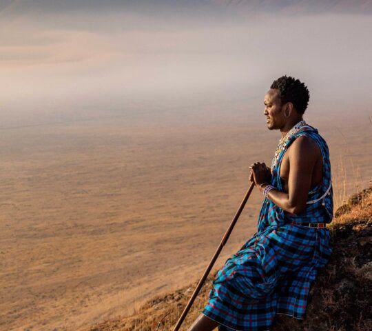 A man in traditional blue clothing sits on a ridge overlooking a misty valley on luxury Ngorongoro Crater safari tours.
