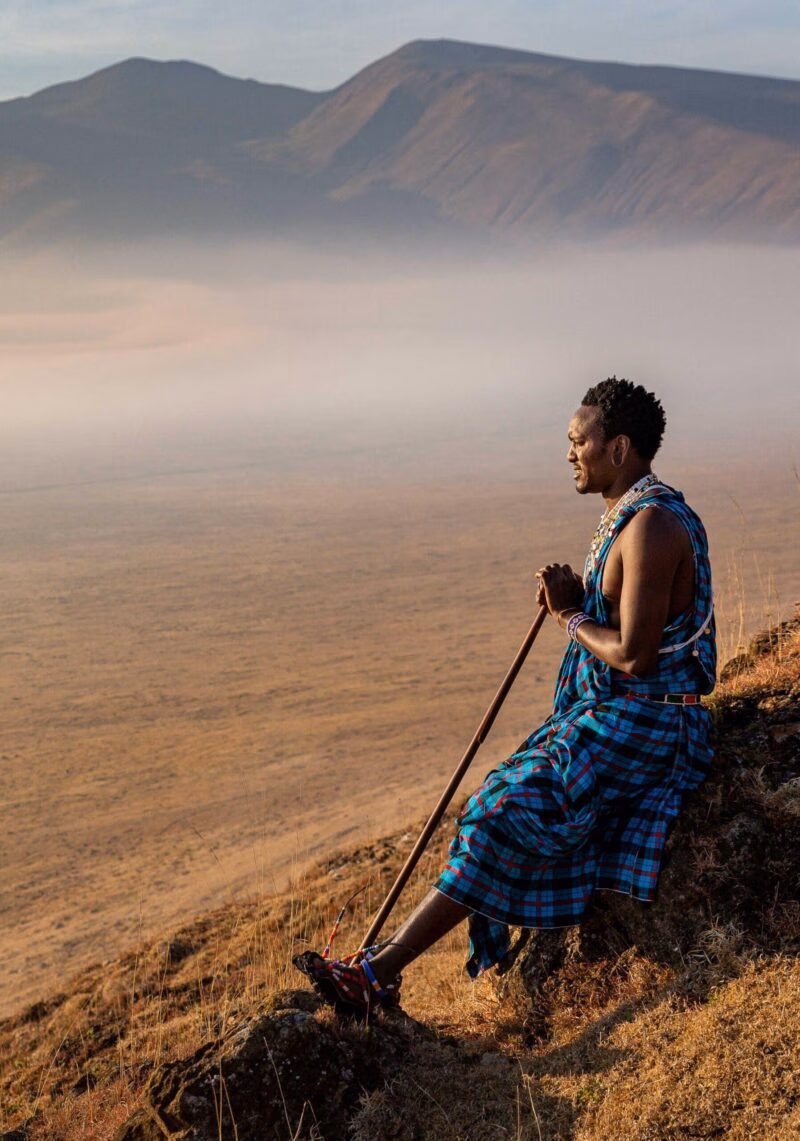 A man in traditional blue clothing sits on a ridge overlooking a misty valley on luxury Ngorongoro Crater safari tours.