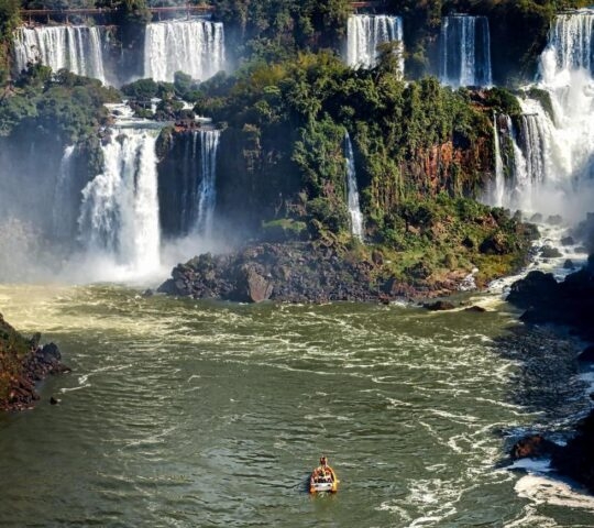 Wide view of cascading waterfalls into a large river with a small boat in the foreground.