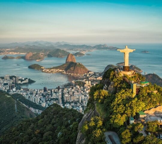 Aerial view of Christ the Redeemer statue overlooking Rio de Janeiro's coastline and Sugarloaf Mountain at dusk.
