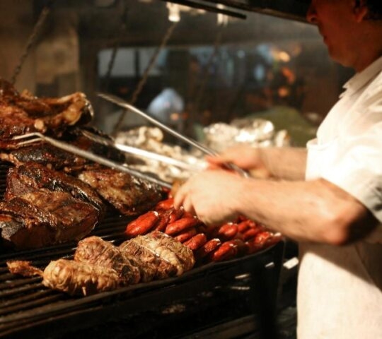 A chef grilling large steaks and red sausages on a metal grate with smoke rising.