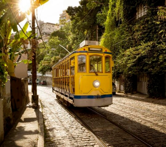 A yellow vintage tram travels down a cobblestone street lined with green trees and buildings in sunlight.