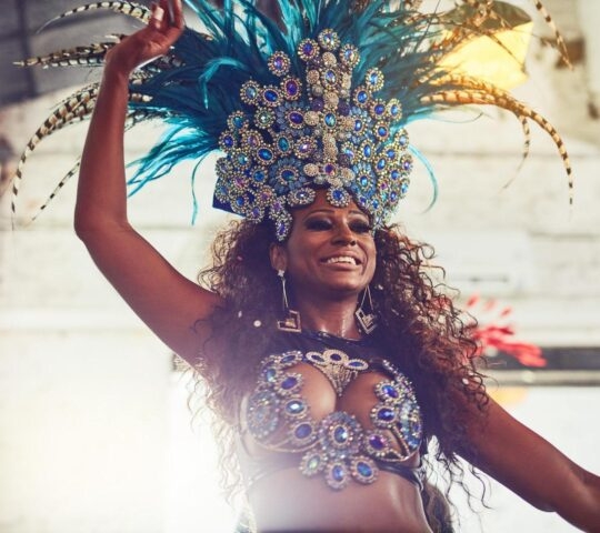 A smiling woman wearing an elaborate blue and silver feathered headdress and jeweled costume performing.